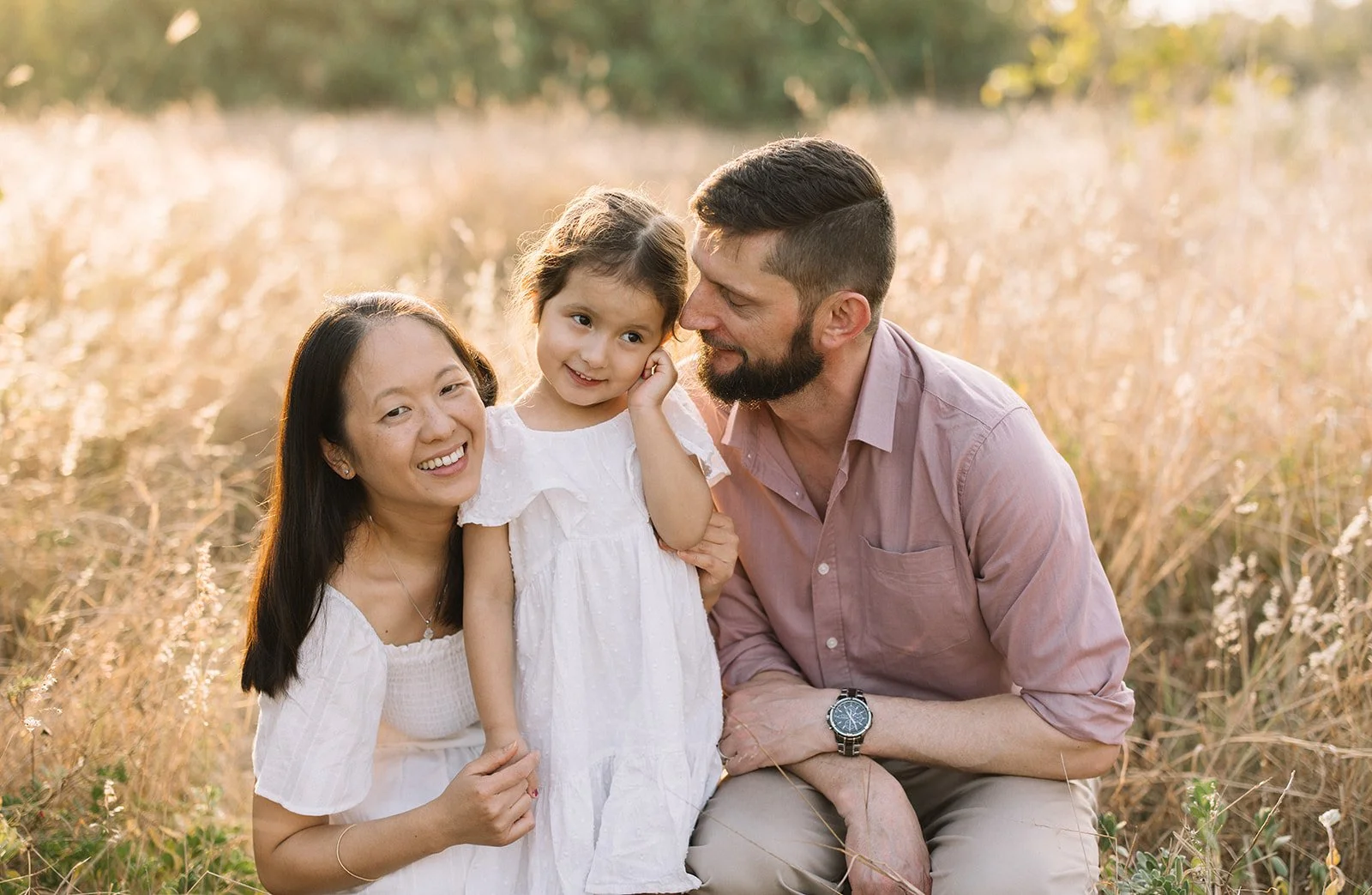 Family of three, a woman, man, and young girl, sitting in a field of tall grass during sunset, smiling and spending time together. Taken by Wylde Folk Studio, Brisbane.