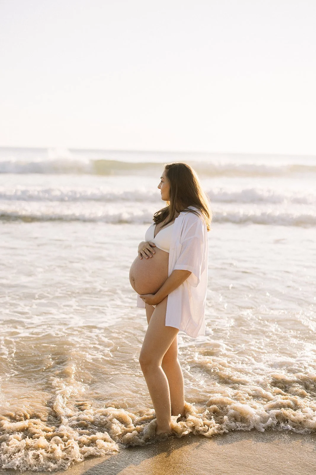 Pregnant woman standing in ocean waves at sunset, wearing a white bra and an open white shirt. Taken by Wylde Folk Studio, Brisbane. Maternity photography.