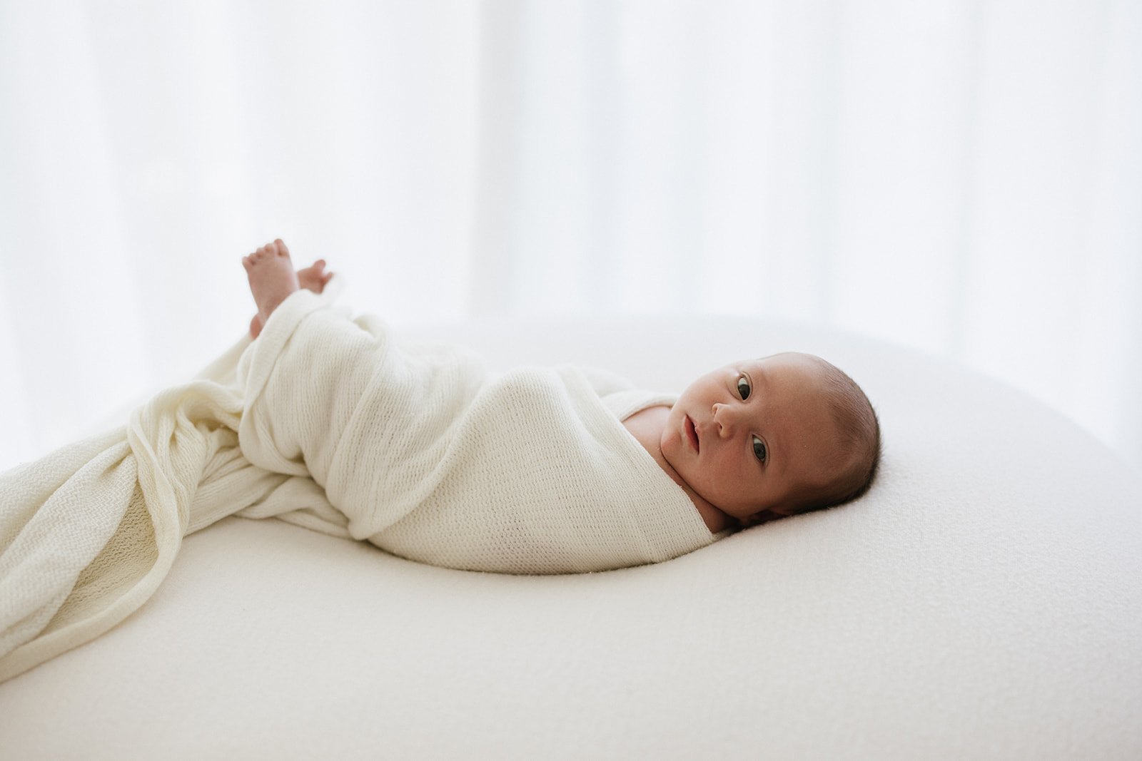 Newborn baby wrapped in white cloth and lying on a white cushion. Baby is looking at camera