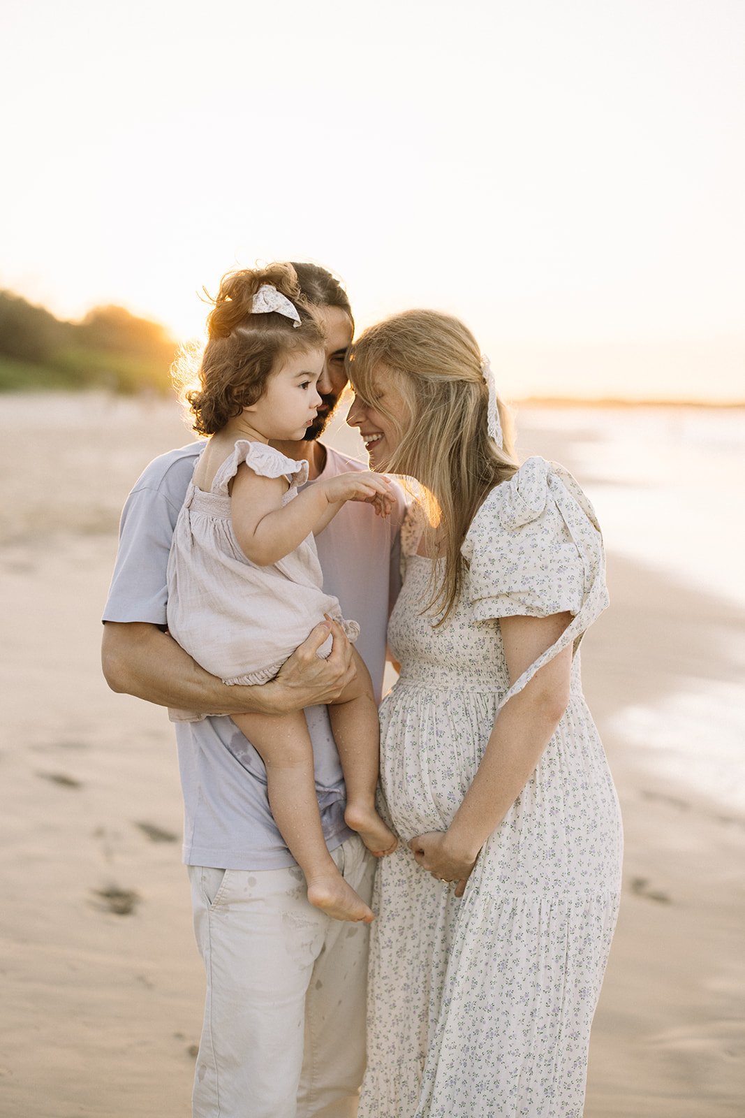 A family consisting of a man, a woman, and a small girl standing on the beach during sunset, touching foreheads and smiling. Taken by Wylde Folk Studio, Brisbane. Maternity photography.