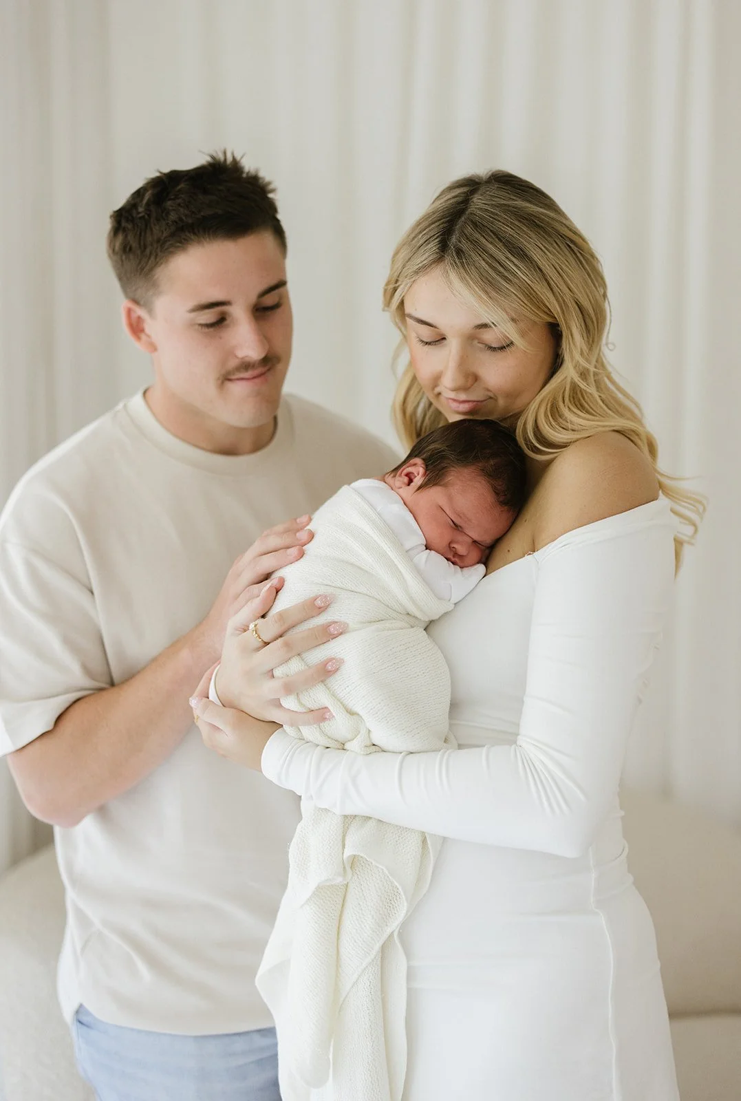 A mother holding a newborn baby wrapped in a white blanket, with a man looking on, in a bright, softly lit room.
