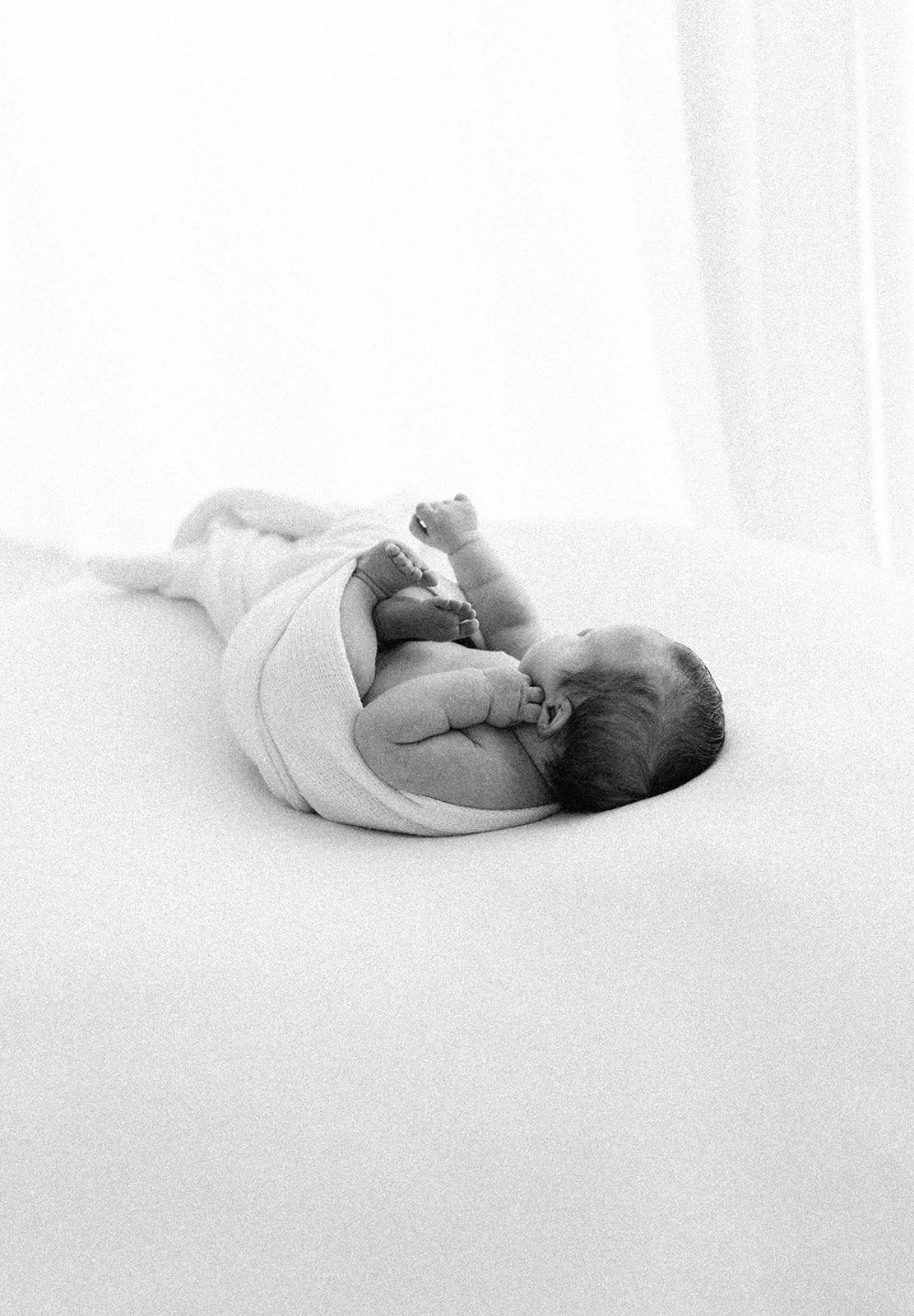 Black and white photo of a newborn baby lying on a soft surface, holding its feet, with a peaceful expression.