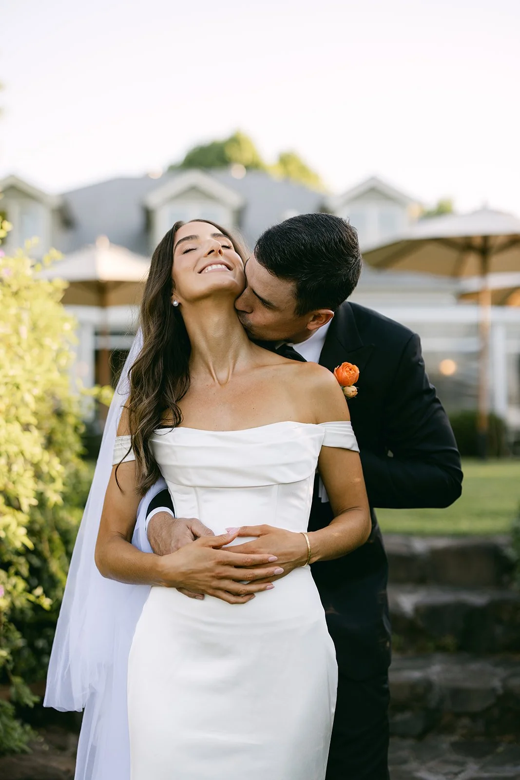 Bride and groom standing in-front of venue. Groom kissing bride's neck.