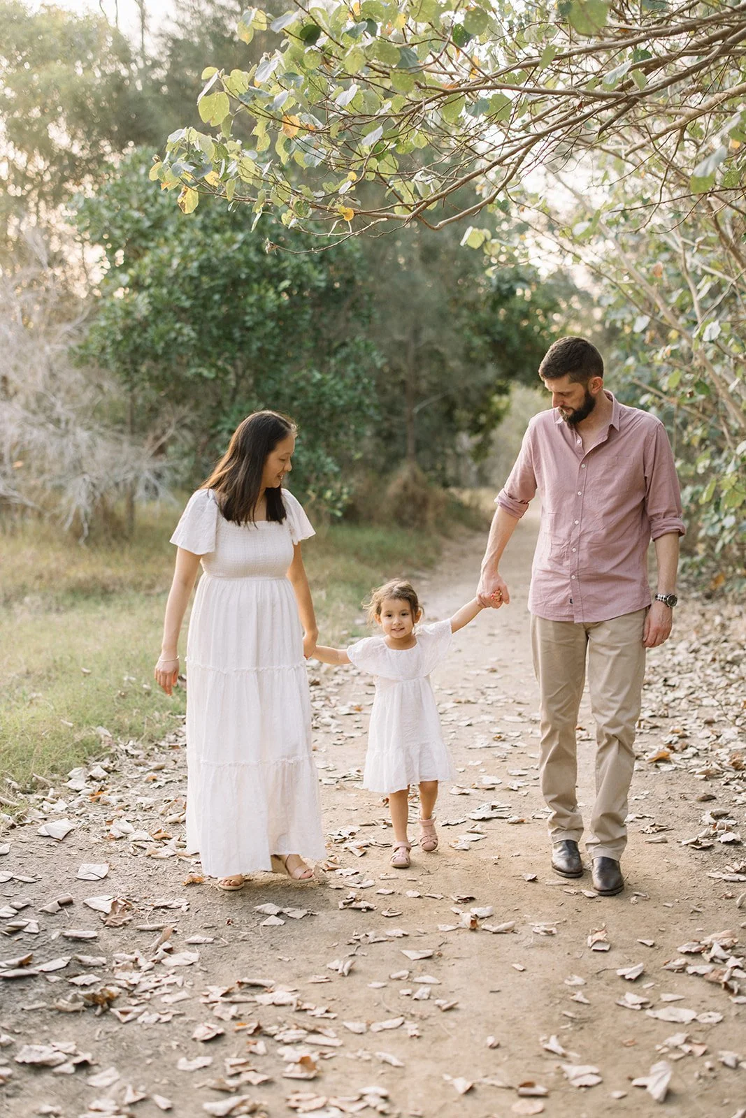 A family of three, a woman, a man, and a young girl, walking hand in hand on a dirt path surrounded by trees and fallen leaves. The woman and girl are dressed in white, while the man wears a light pink shirt and beige pants. The scene is bathed in wa