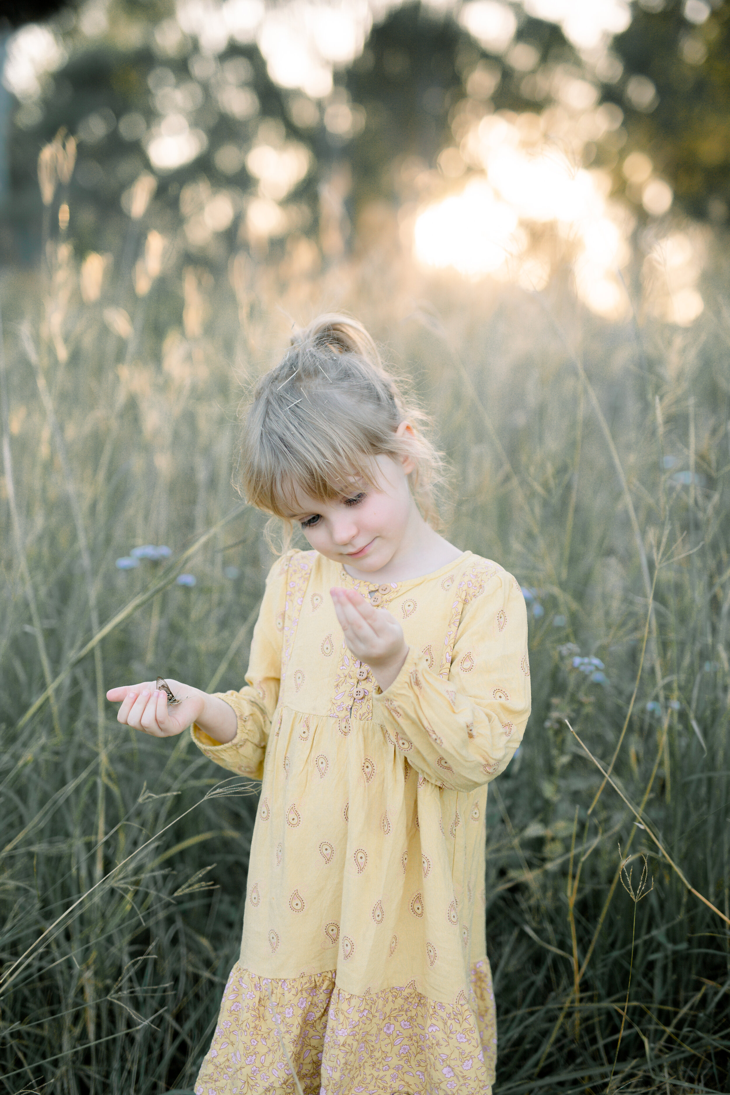  Little girl holding a butterfly in her hand. The girl is in a grassy field wearing a yellow dress. 