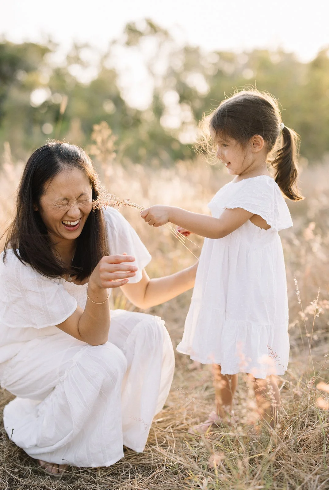 A mother and a daughter sharing a playful moment outdoors in a field, both wearing white dresses, with the girl tickling the woman with a piece of tall grass. Taken by Wylde Folk Studio, Brisbane. Family photographer.