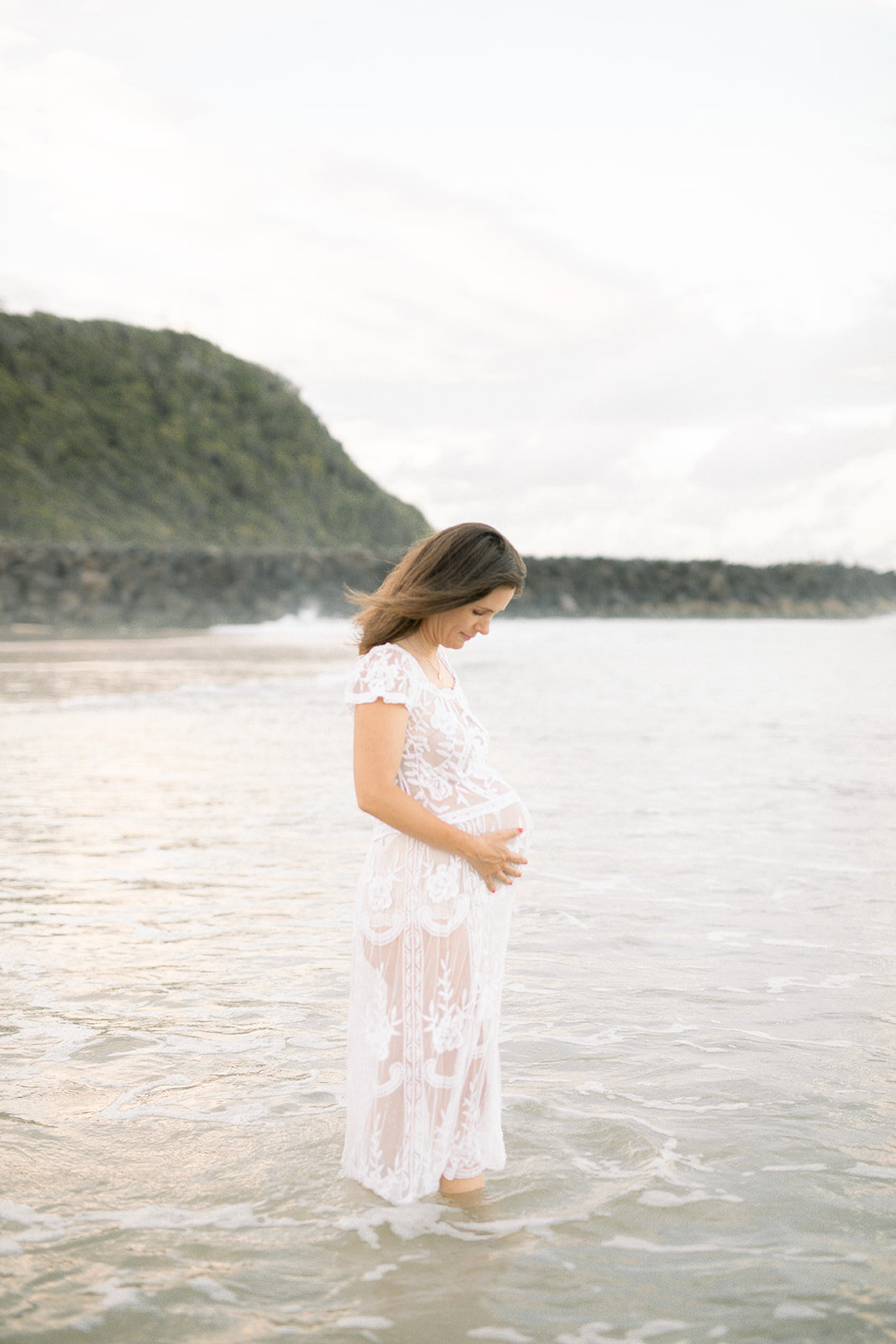 Mother standing in the water holding her baby bump in a white gown 