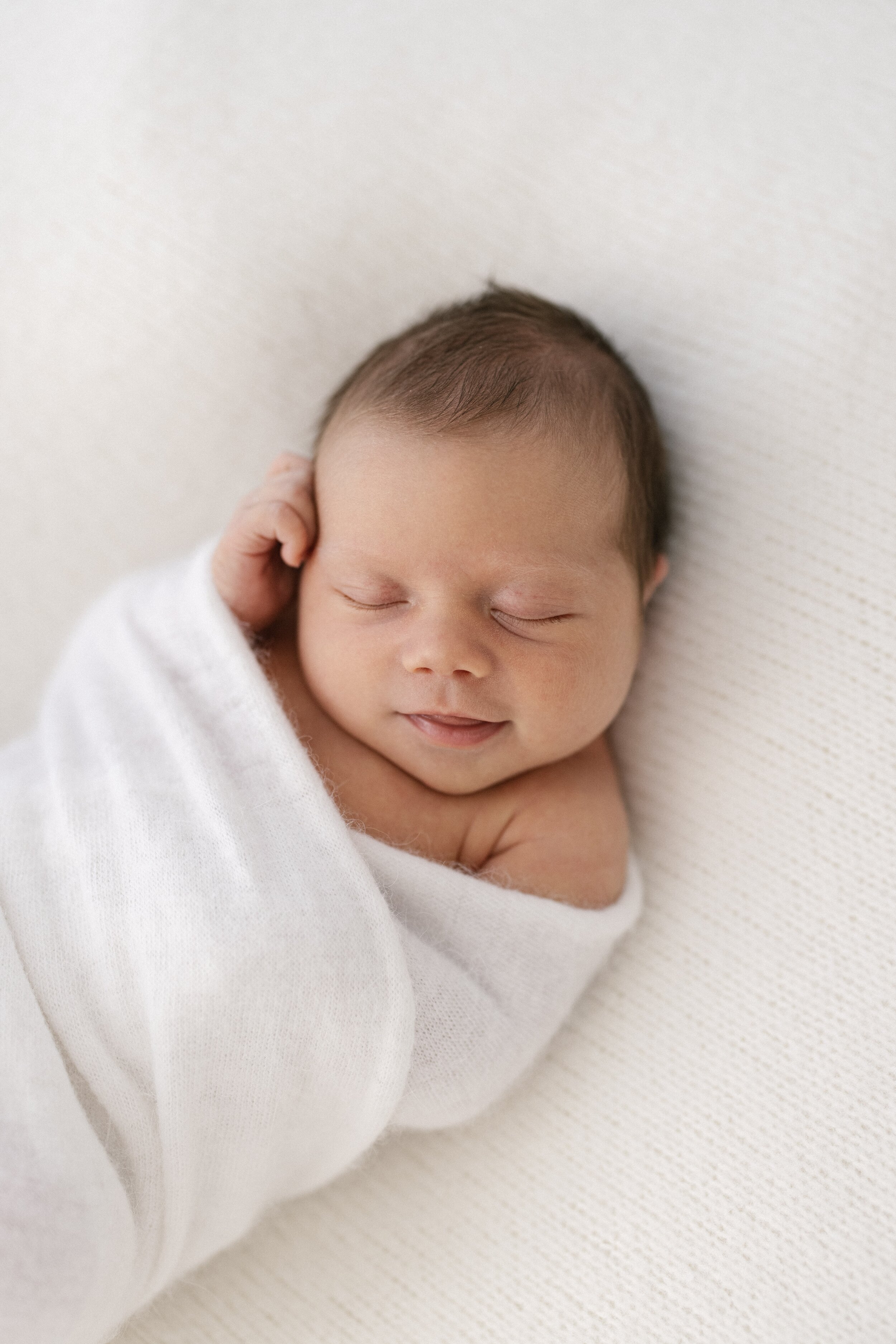 newborn baby smiling with dark hair wrapped in fluffy white wrap 