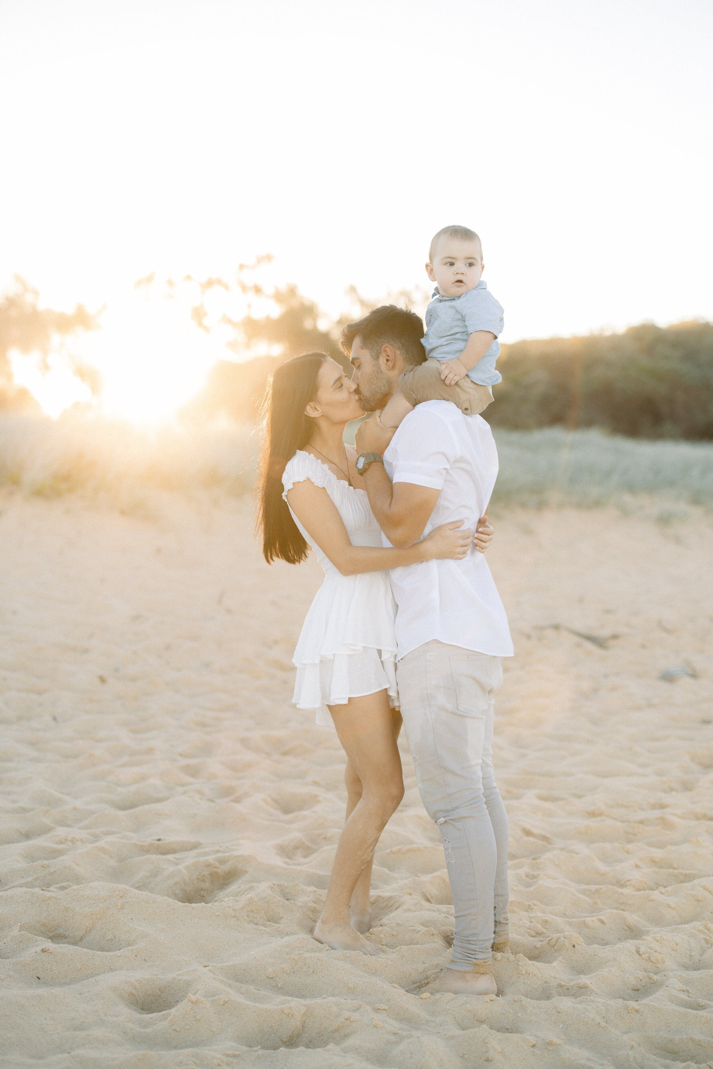 Couple kissing on the beach with their baby on Dad's shoulders