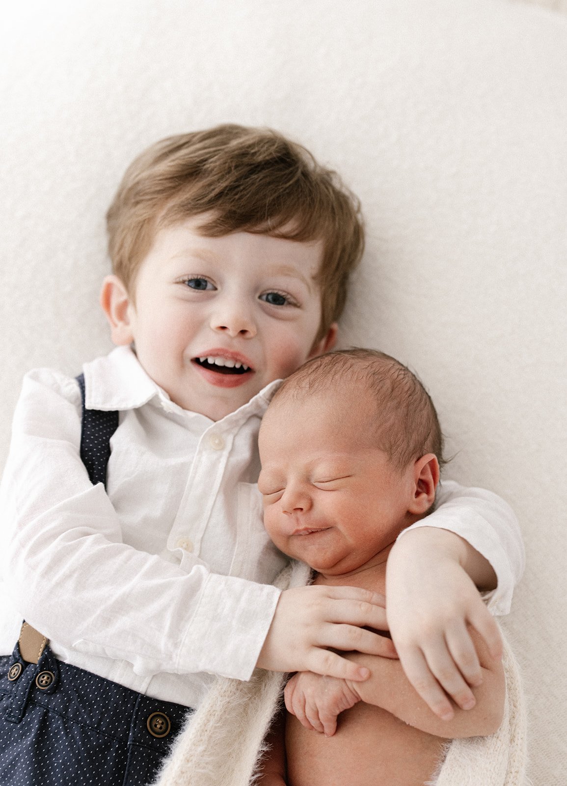 a big brother wearing blue suspenders cuddles his new newborn baby brother