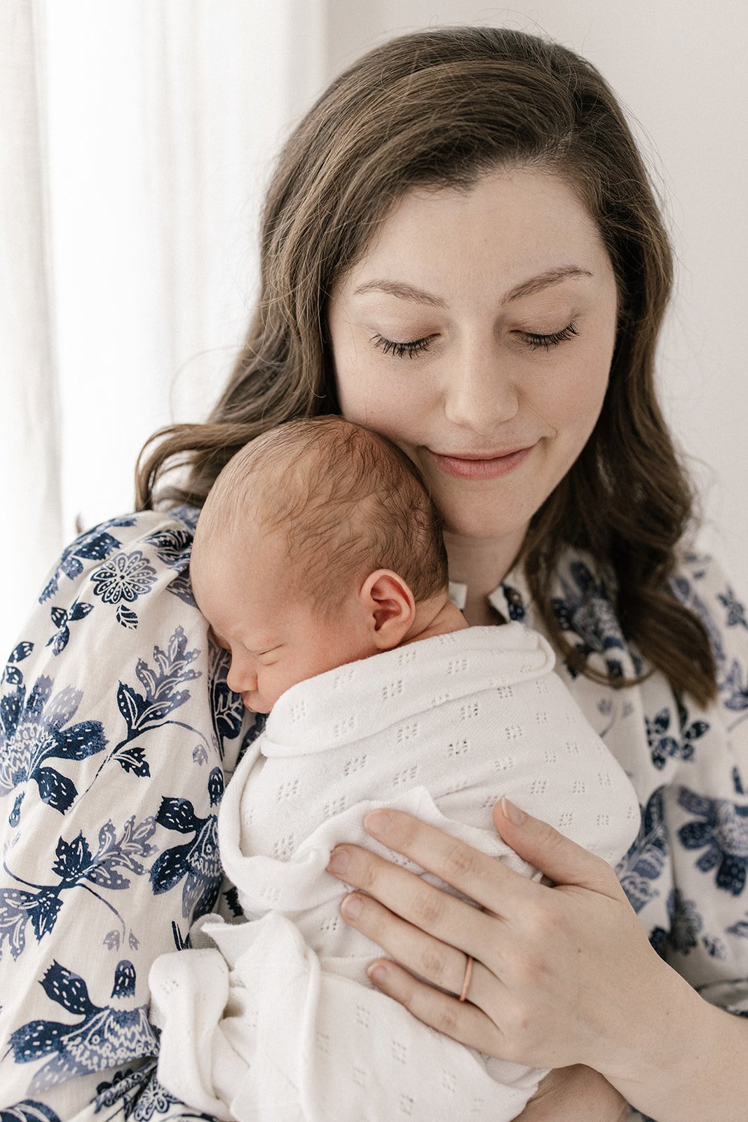 A mother with loosely curled dark hair wears a blue floral patterned dress with cuddling her newborn baby who is wrapped in a white blanket