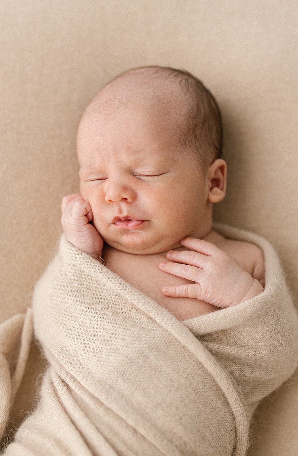 A newborn with his hand pressed against his cheek is wrapped in a latte coloured wrap