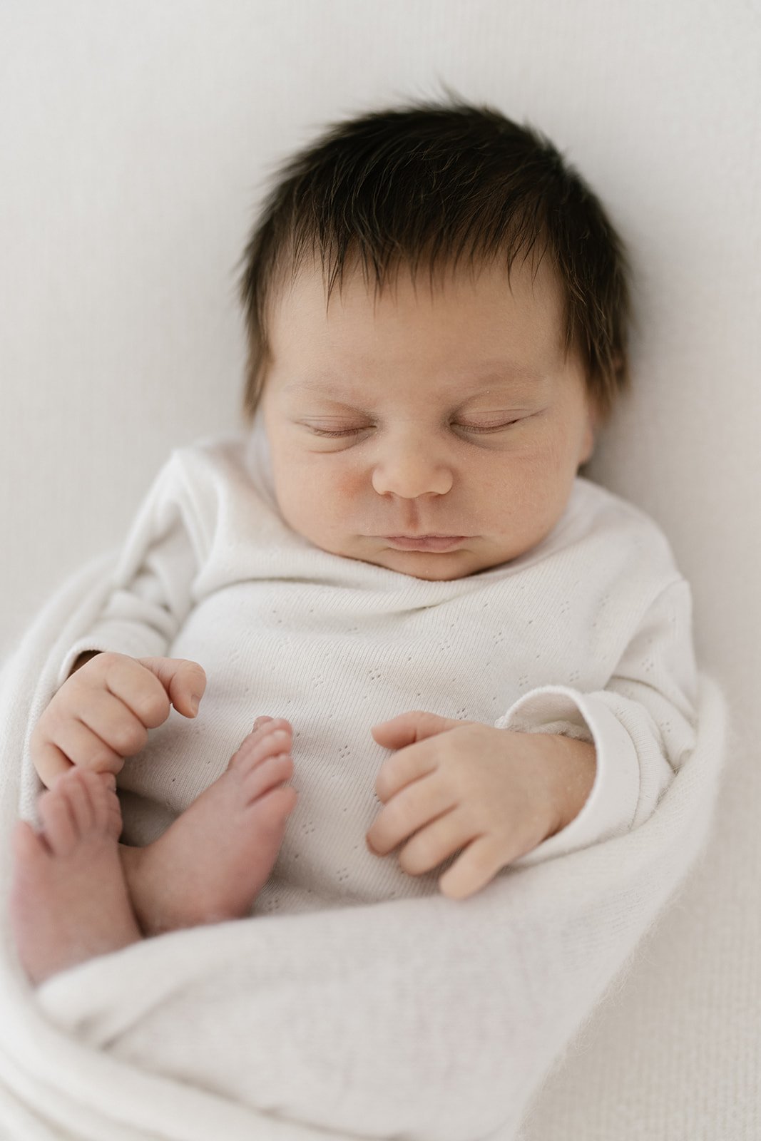 a newborn baby with thick dark hair is sleeping and wrapped in a fluffy white wrap