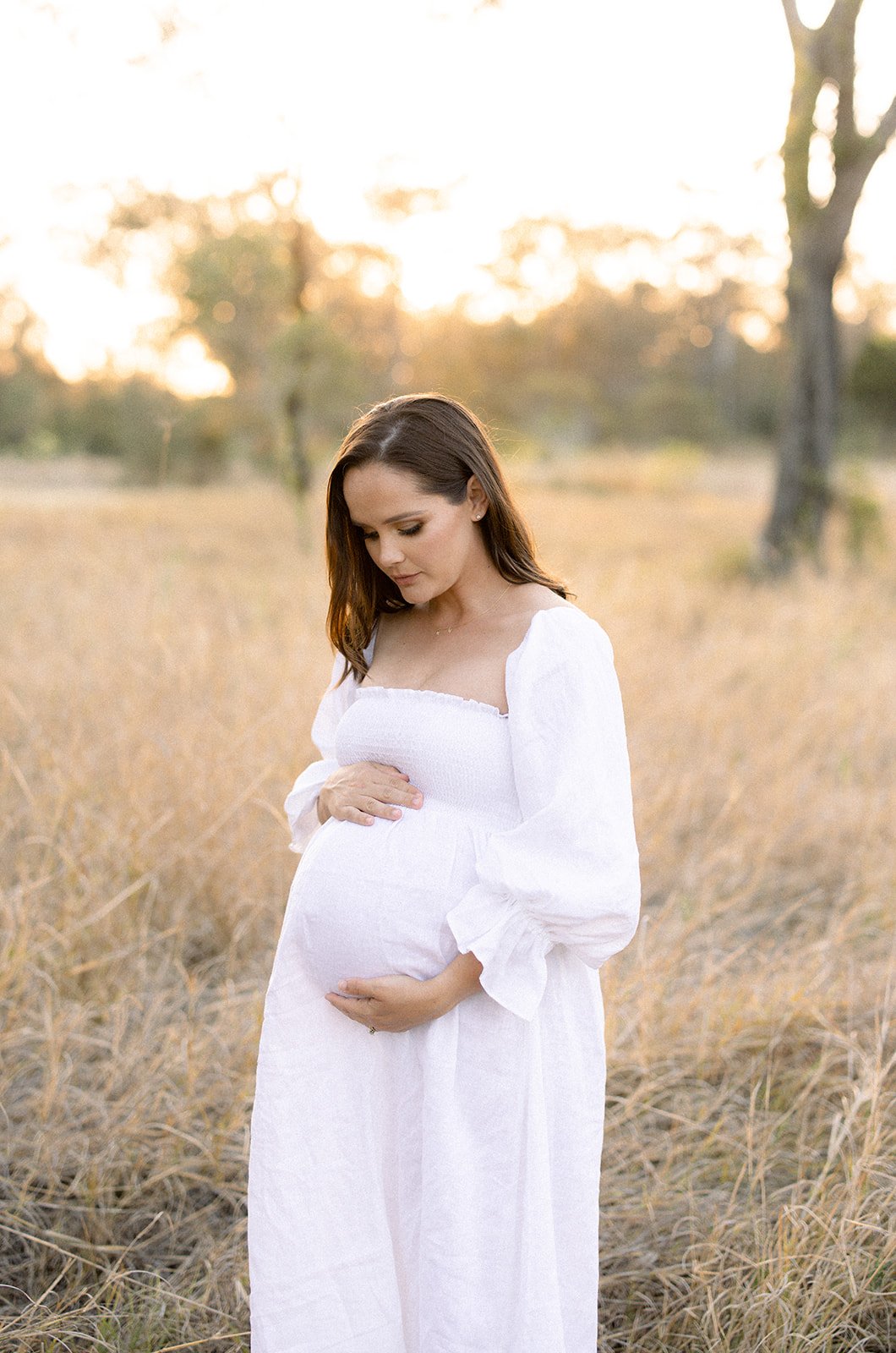 Expectant mother wearing a white dress holds her baby belly in an open field