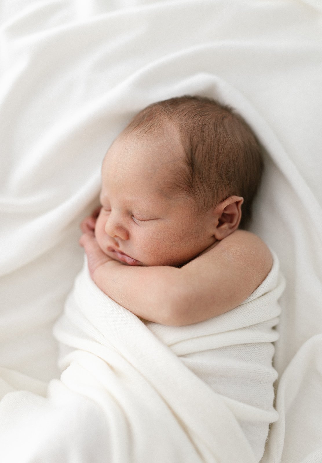 newborn baby with dark hair cuddled on its side with white ruffled blankets surrounding him