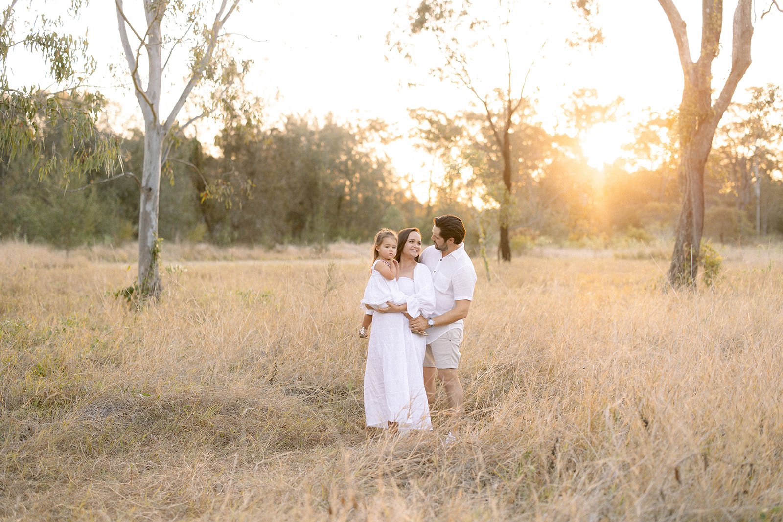 mother sits her toddler on her growing baby bump while dad cuddles from behind in a sunset field