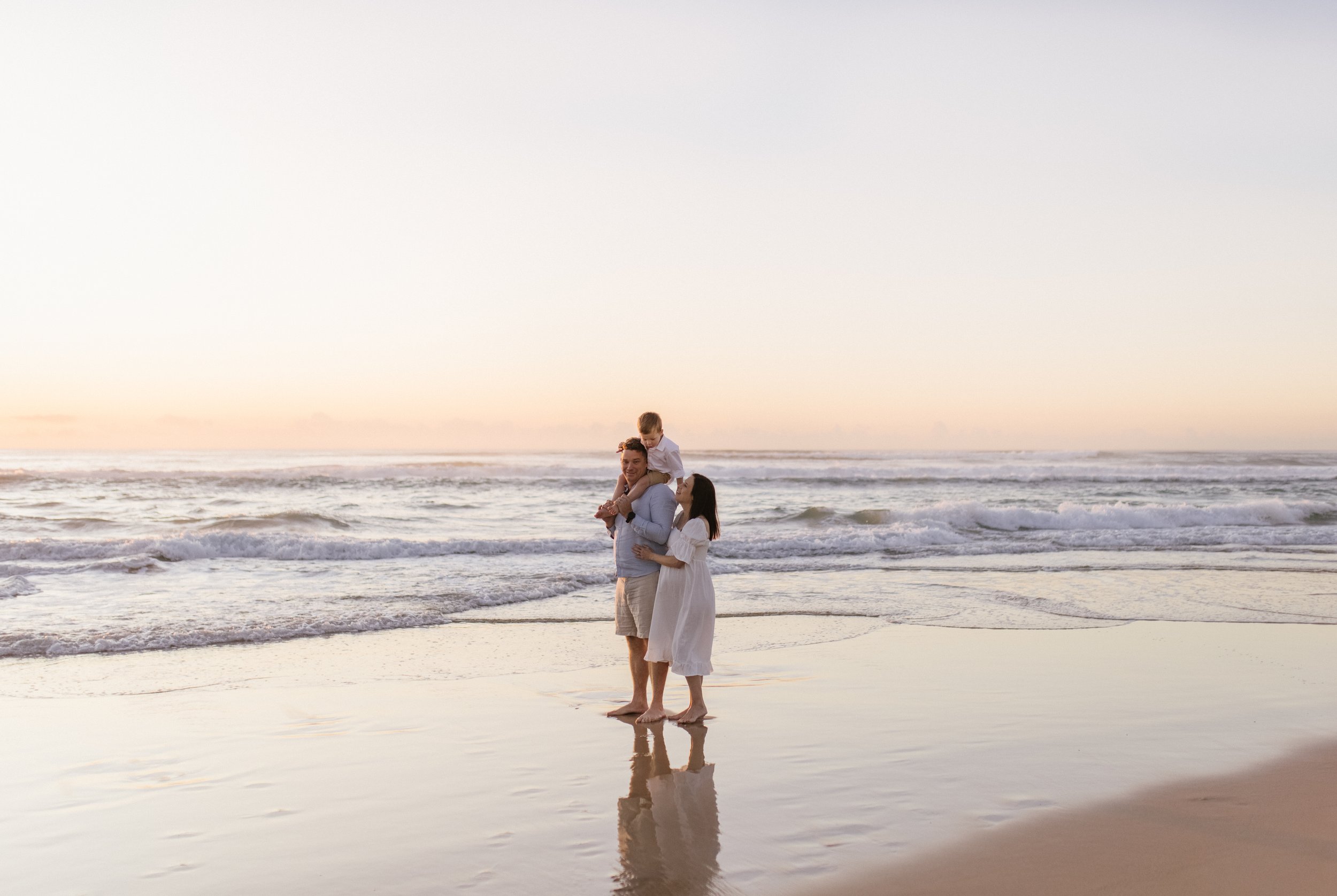 A family cuddle together at sunrise on the beach, pink skies and a pregnant belly can be seen