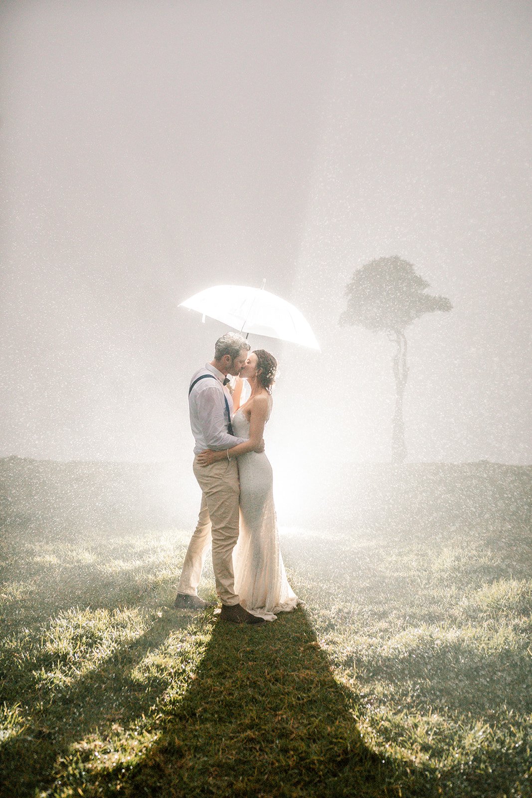A bride and groom stand under a clear umbrella on a rainy wedding day in Maleny