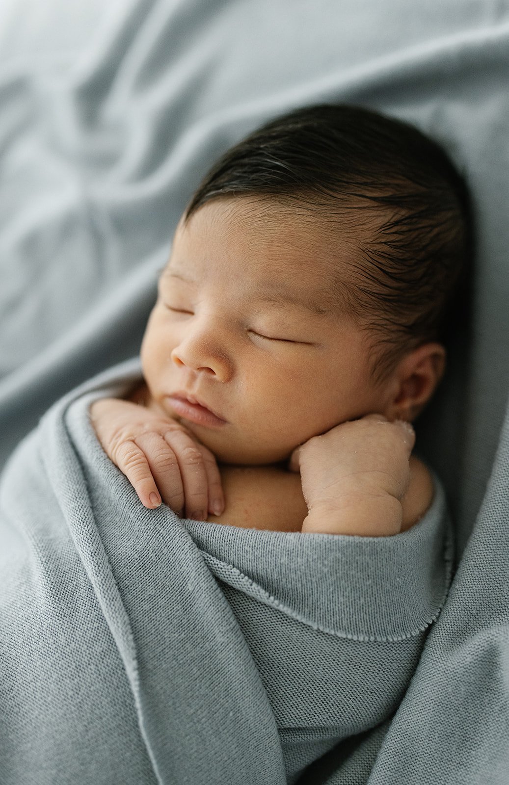 newborn baby boy photographed with dark thick hair and wrapped in a blue blanket