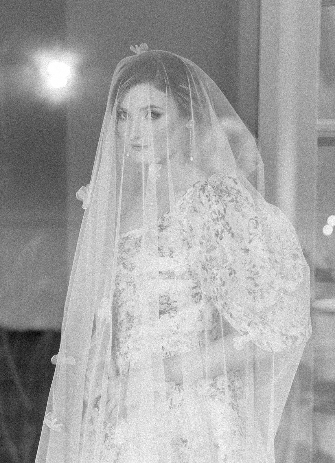 A bride is draped in a veil as she stares straight at the camera. The veil features tiny flowers dotted through out. The photo is in black and white