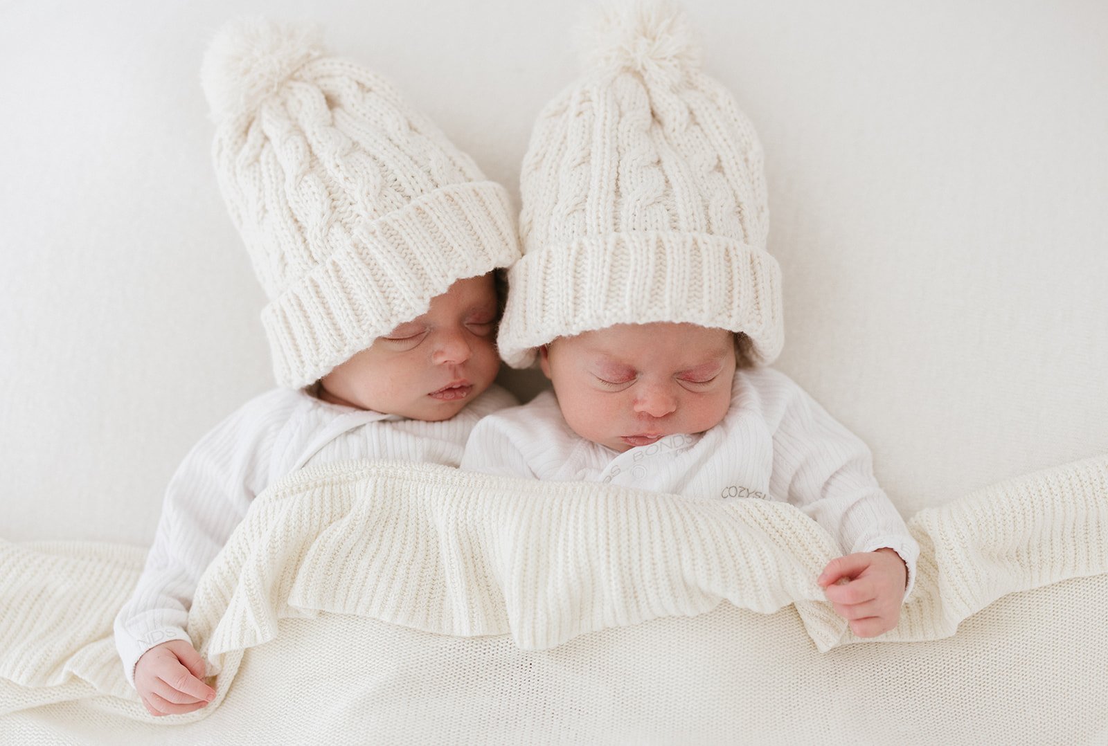  newborn baby twins photographed in white knit beanies cuddled in a white frill blanket  