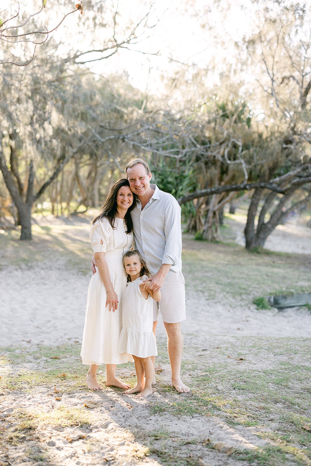 a mum, dad and their young daughter cuddle up under the beach trees and smile for a family photo