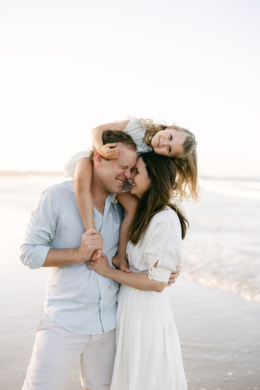 a young girl with curly blonde hair in sitting on her dad shoulders. Mum and dad are cuddled close together and the little girl leans her head over to sit on the top of mums head.