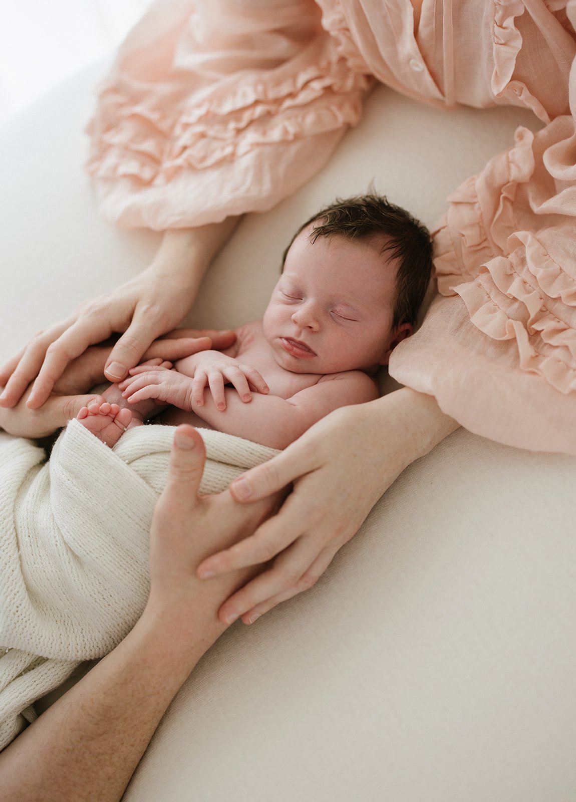 Mum and dads arms are cuddled into their newborn baby girl. Mum is wearing a pink ruffled dress