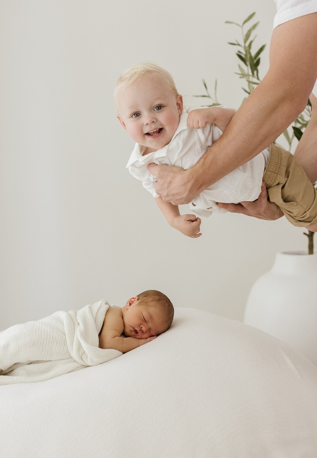 A toddler is flying over their newborn baby sibling in dads arms. He is giving the photographer a big cheesy grin. 