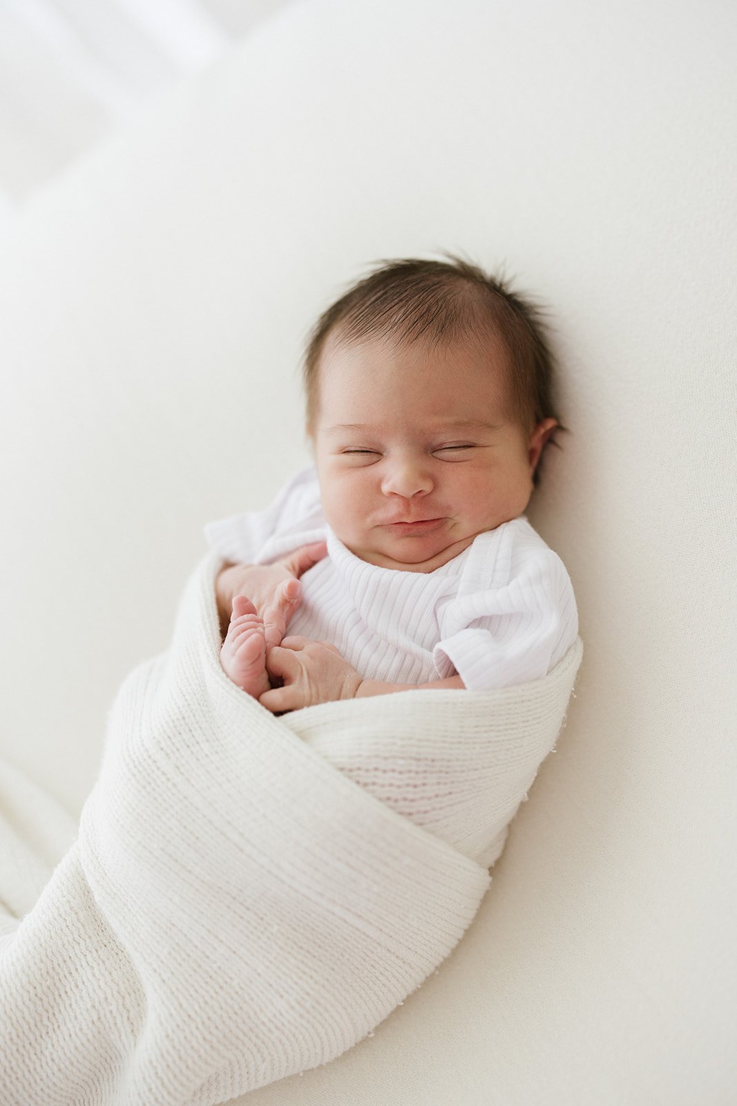  a newborn baby with dark hair is photographed in a white wrap. The baby has a cute squishy face and is smiling