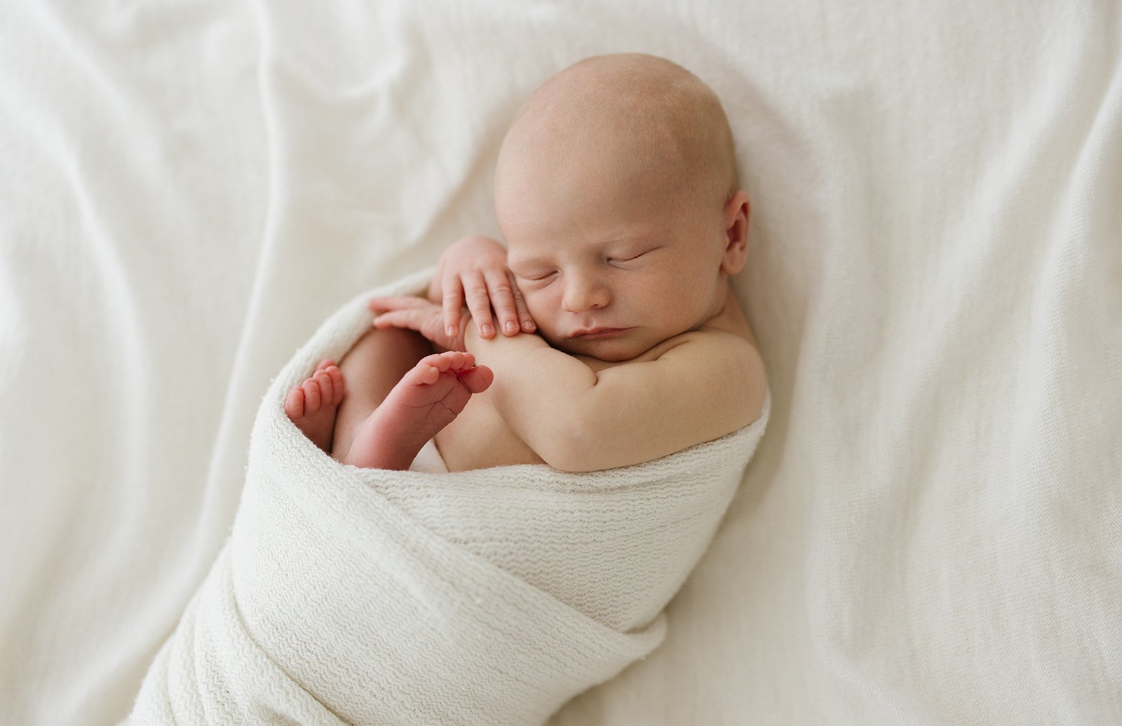 A newborn baby boy is wrapped in a white blanket. Their little feet poking out the top