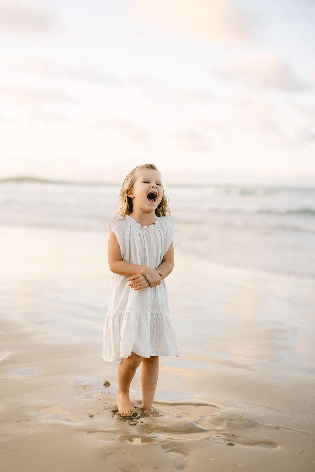 a young girl in a white dress is standing on a Sunshine Coast beach and laughing loudly into the air