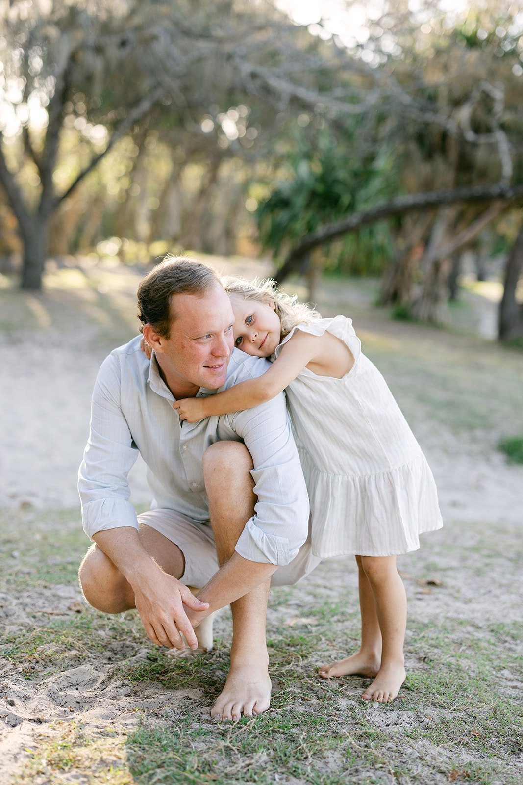 a little girl with blonde curls leans on her dads shoulder as she cuddles into him at Noosa main beach during a family photography session