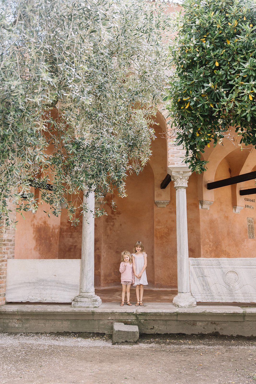 two little girls in pretty pink dresses stand under a giant olive tree in Venice Italy 