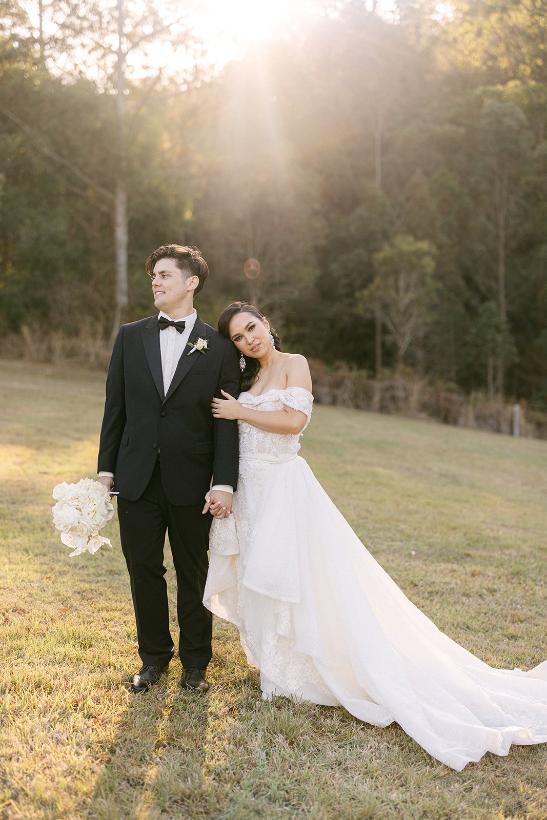 A bride in a long white gown leans on her grooms shoulder. A golden sunset is behind them