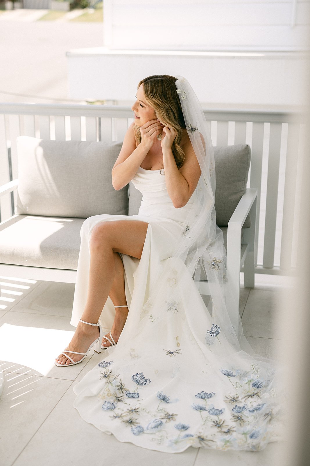 A Brisbane bride is sitting on a bench seat while she puts in her earrings. She is wearing a flowing veil with little blue flowers that feature on the bottom