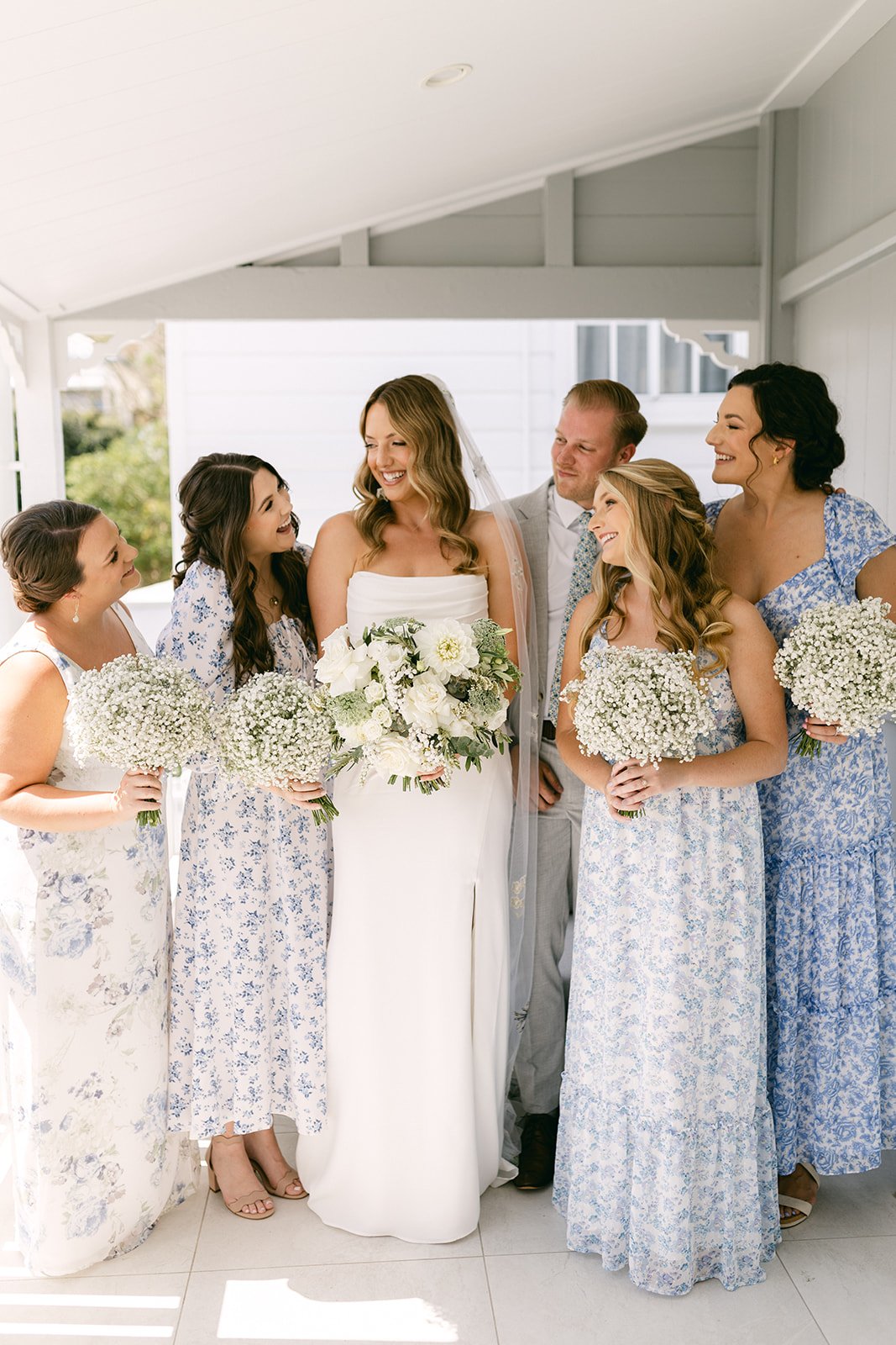 A bride wearing a strapless white dress is surrounded by her bridesmaids and bridesmaid. The girls are wearing mix match blue and white floral dresses