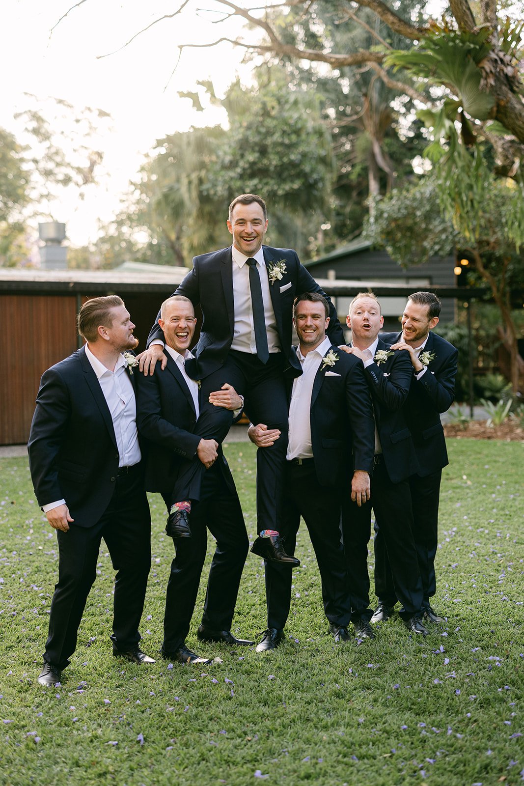 A groom sitting on top of his groomsman's shoulders. They boys are laughing and throwing him up in the air