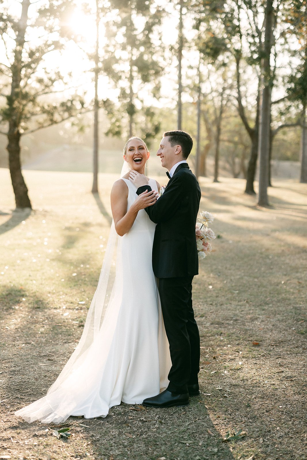 A bride and groom are drenched in afternoon golden sunlight and they laugh. The grooms hand is sitting gently on the brides cheek