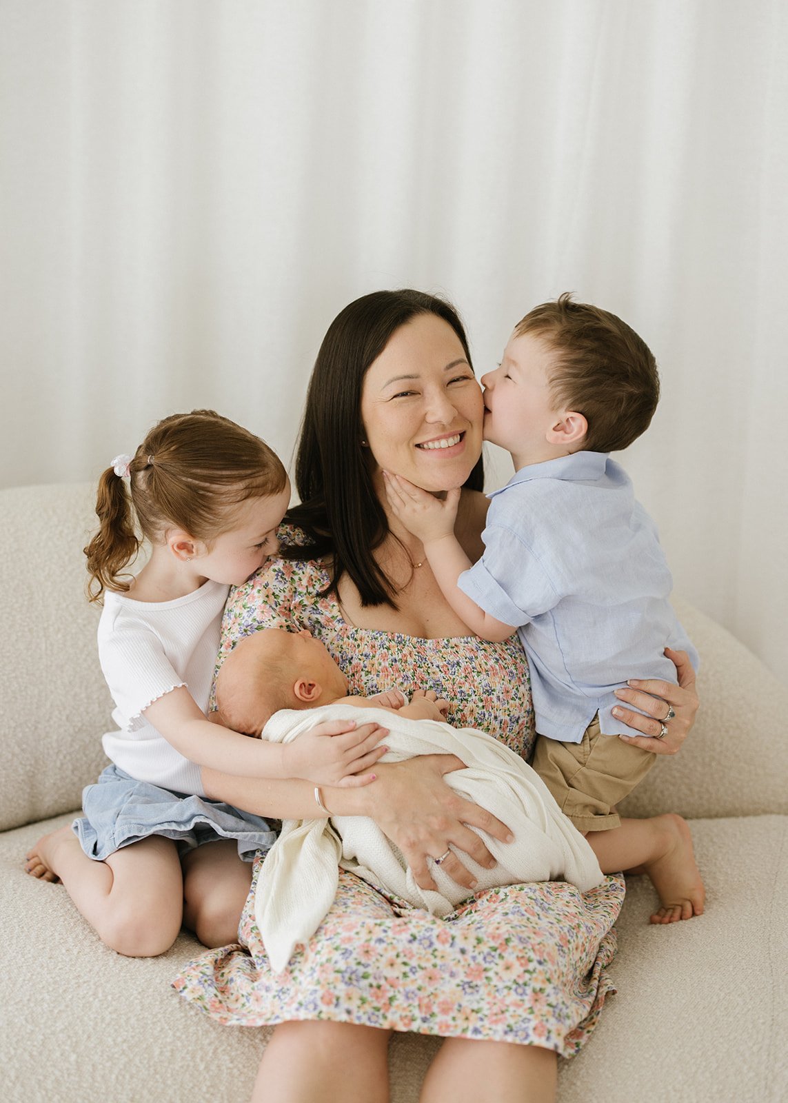 A woman surrounded by three young children, one kissing her on the cheek, one cuddling her on her lap, and one kissing her on the cheek, all on a beige couch in front of a neutral background.