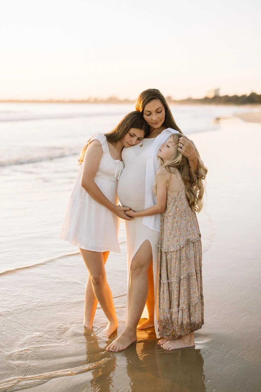 A pregnant woman at the beach with two young girls, all embracing each other tenderly during sunset. Taken by Wylde Folk Studio, Brisbane. Maternity photographer.