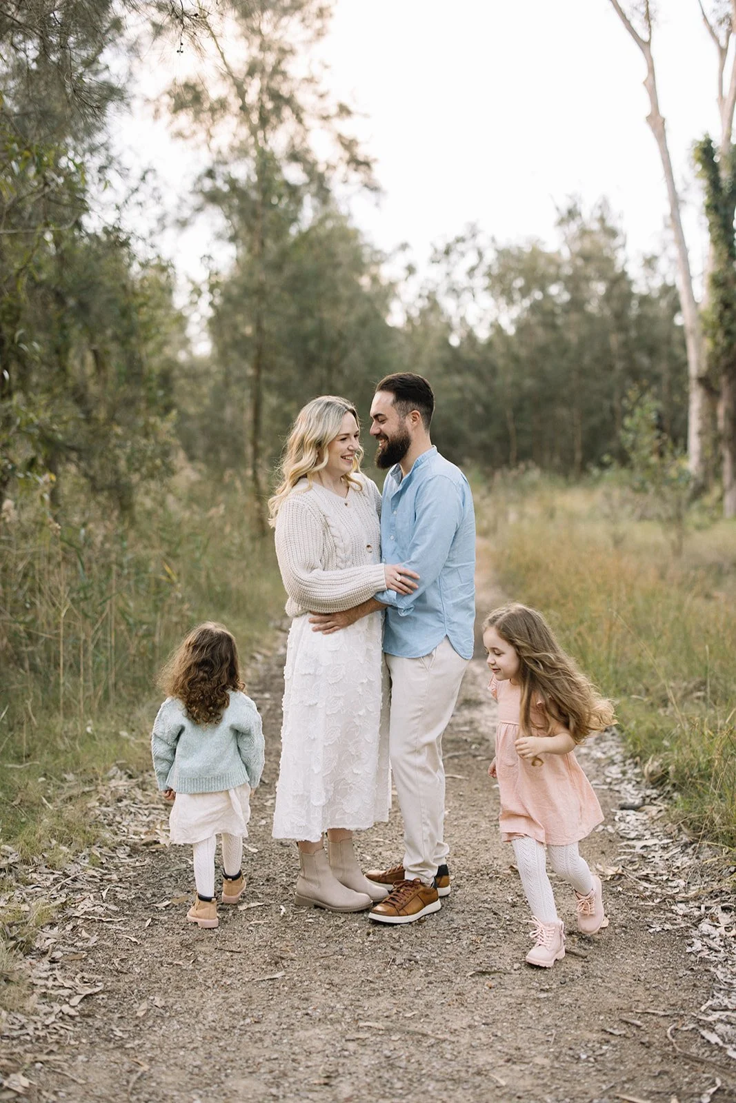 A family of four standing on a dirt path in a wooded area, smiling and looking at each other, with the parents embracing and the two young girls playing around them. Taken by Wylde Folk Studio, Brisbane. Family photography.
