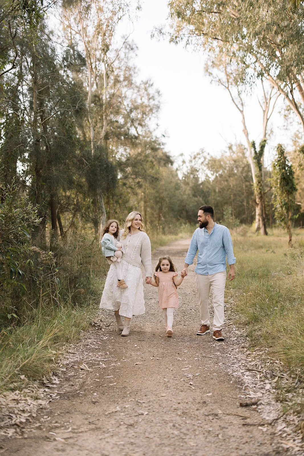 A family of four walking on a dirt path in a wooded area during daylight, holding hands and smiling, with tall trees around them. Taken by Wylde Folk Studio, Brisbane. Family photographer.