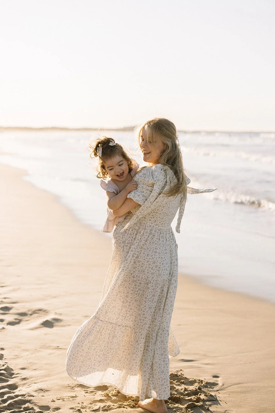 A woman holding a young girl on a beach at sunset, both smiling and enjoying their time together. Taken by Wylde Folk Studio, Brisbane. Maternity photographer.