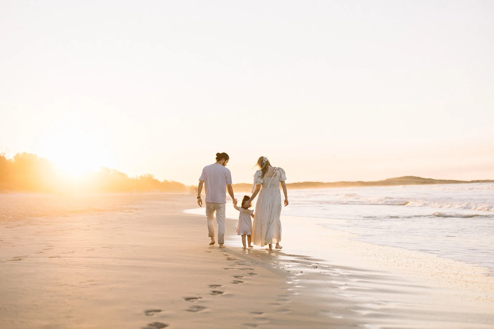 A family of four holding hands walking on the beach at sunset, with the ocean waves and shoreline in the background. Taken by Wylde Folk Studio, Brisbane. Maternity photographer.