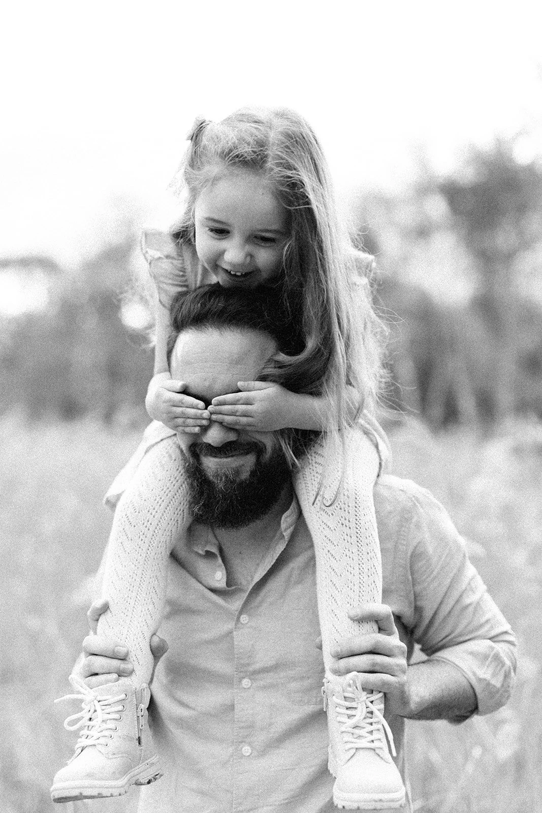 A man giving a piggyback ride to a young girl, who is covering his eyes with her hands and smiling. Black and white photo sit gunwood outdoors. Taken by Wylde Folk Studio, Brisbane. Family photographer.