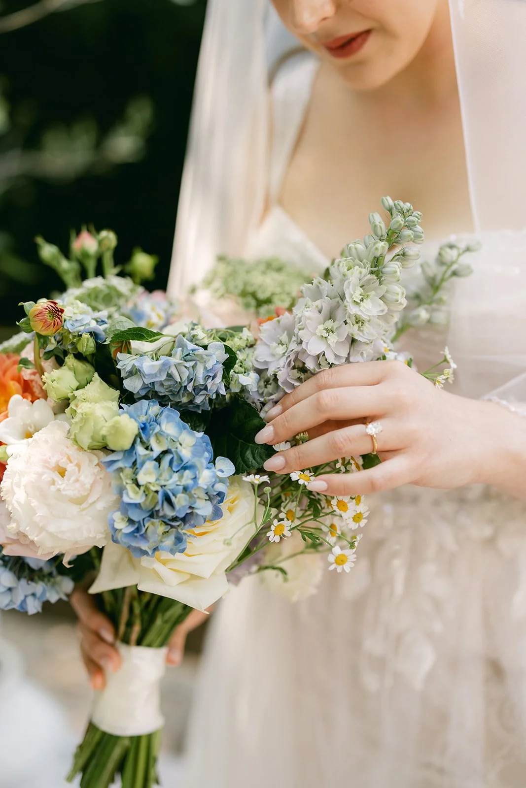Close-up of a bride holding a bouquet of colorful flowers, wearing a wedding dress and a diamond ring.