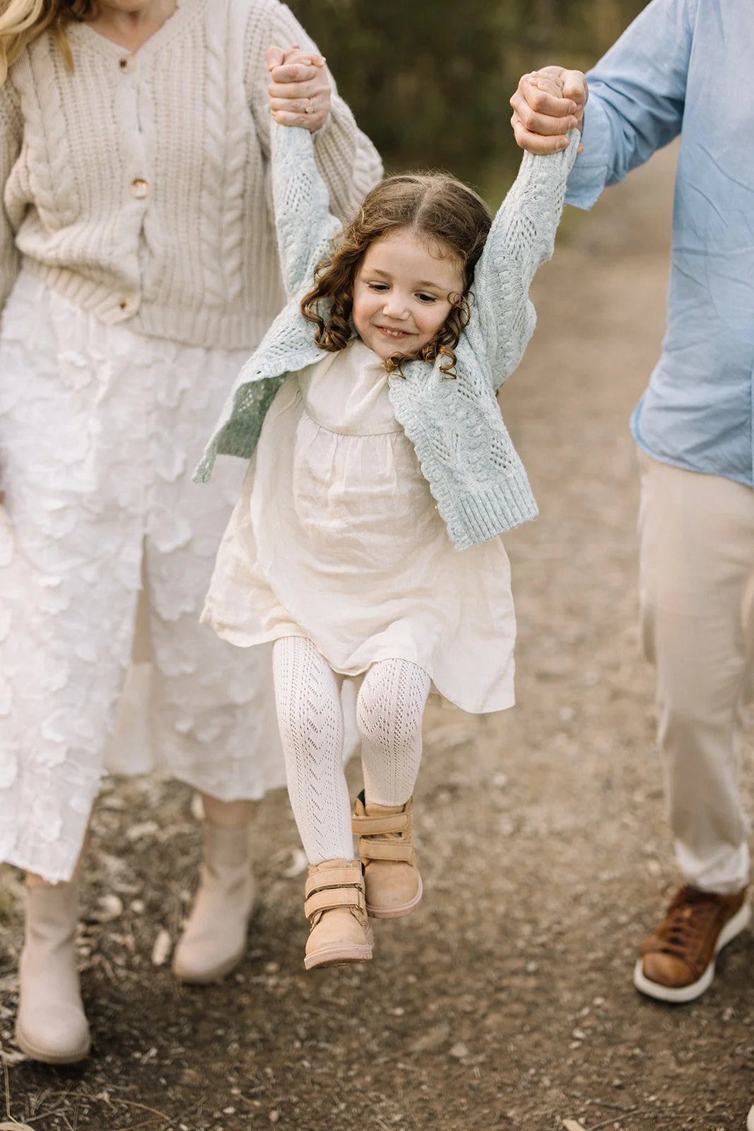Young girl being lifted by two adults, smiling, outdoors on a dirt path. Taken by Wylde Folk Studio, Brisbane. Family photography.