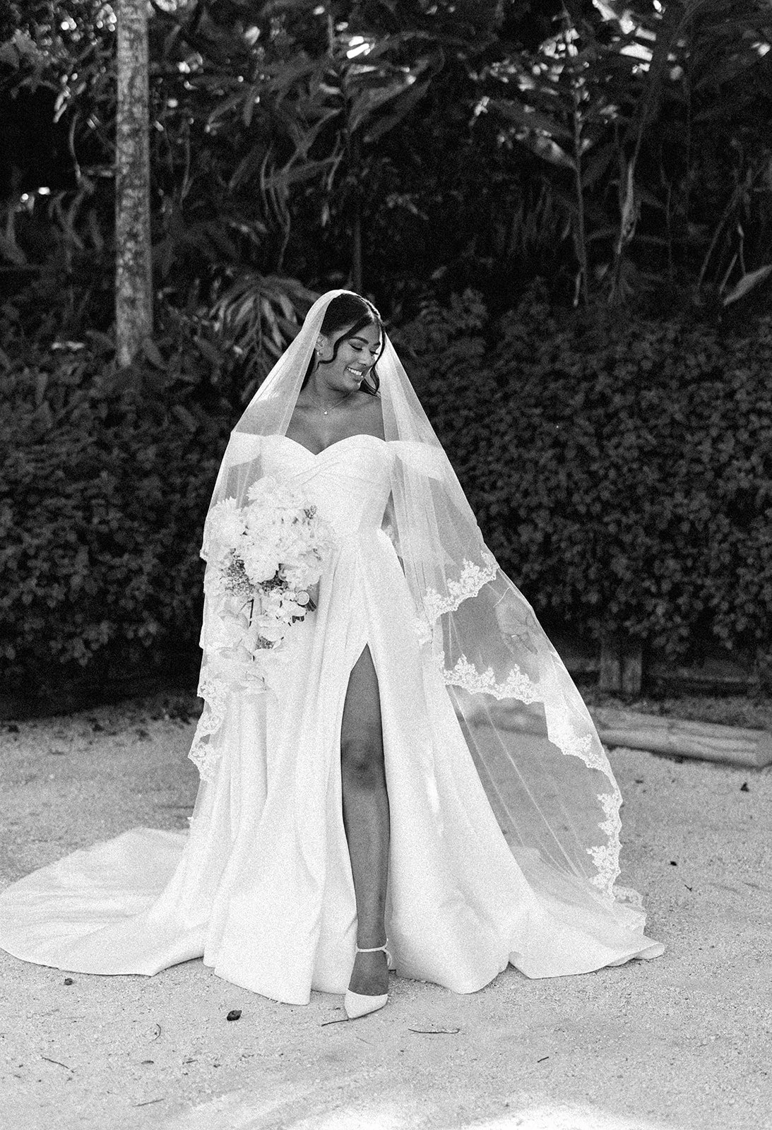 A bride in a wedding gown and veil holding a bouquet, smiling and looking down, outdoors with trees and bushes in the background.