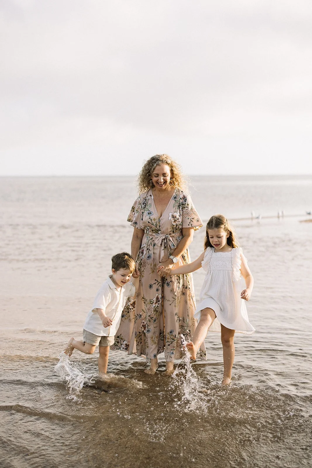 A woman with curly blonde hair and two children playing in the shallow ocean water at the beach, holding hands and splashing. Taken by Wylde Folk Studio, Brisbane. Family photography.